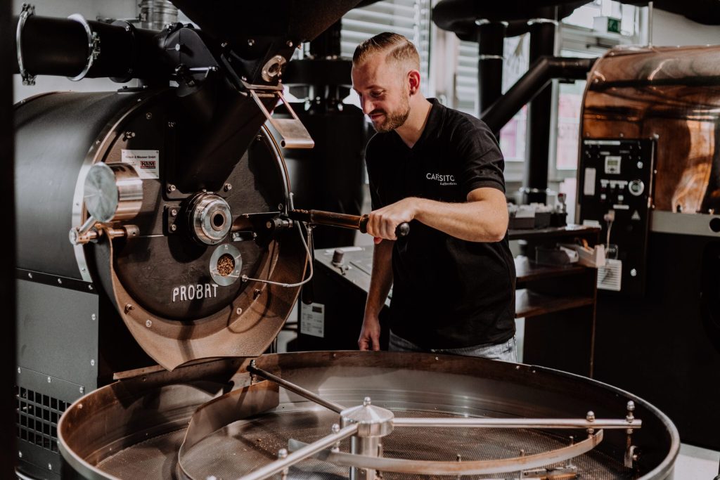 Der Röster steht an einem Trommelröster in der Kaffeerösterei im Allgäu und prüft mit dem Probenzieher, ob der Kaffee fertig ist.